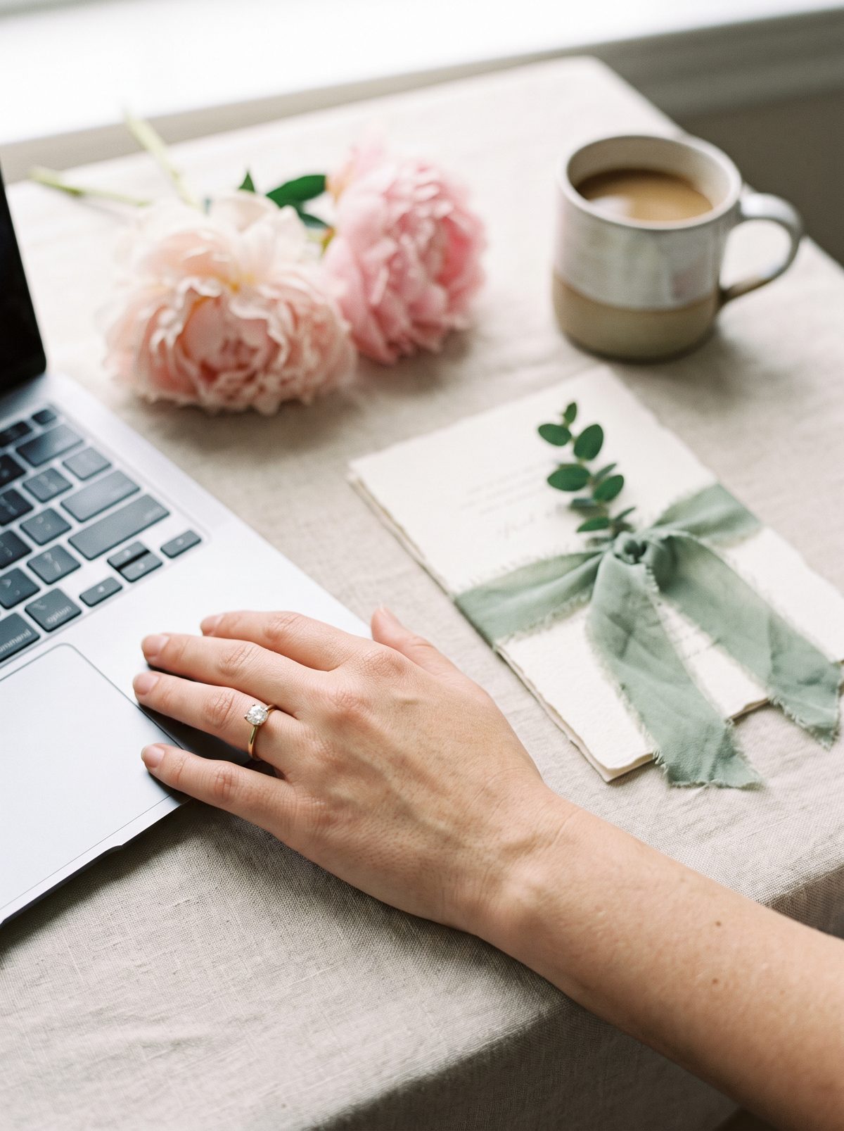 A hand with an engagement ring on a laptop next to a wedding invitation and peonies